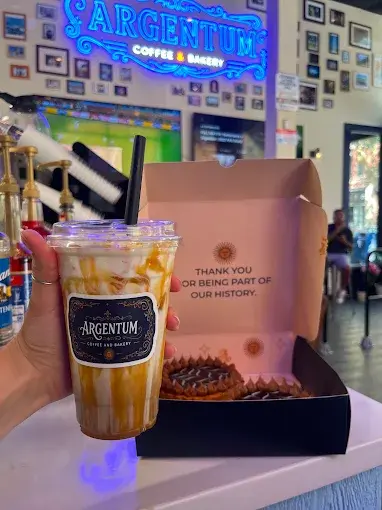 A customer holding an Argentum iced coffee next to a pink pastry box in the shop