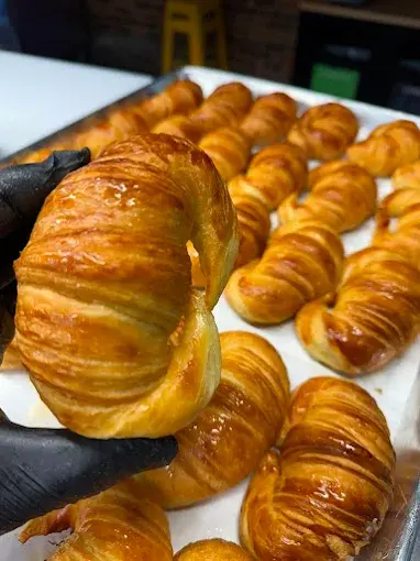 A baker lifting a fresh medialuna from a full tray behind the counter